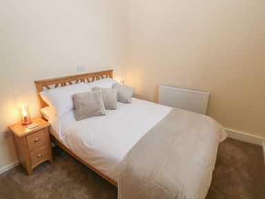 A bedroom with a wooden bed frame white bedding beige pillows and blanket beside nightstand with lamp at Tyddyn Sianel - Derw in Llanystumdwy near Criccieth