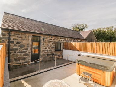 An outdoor patio area with a stone building a hot tub and wooden fencing at Tyddyn Sianel - Derw in Llanystumdwy near Criccieth