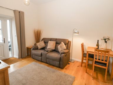 A living room with a brown sofa and three cushions a wooden table with two chairs a floor lamp and a vase with flowers at Tyddyn Sianel - Derw in Llanystumdwy near Criccieth