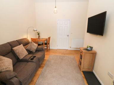 A living room with a brown sofa, wooden dining table with chairs, wall-mounted TV, and wooden cabinet at Tyddyn Sianel - Derw in Llanystumdwy near Criccieth