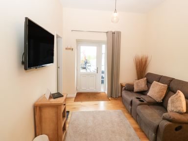 A living room with a gray sofa, wall-mounted TV, wooden cabinet, rug, and a door with a curtain at Tyddyn Sianel - Derw in Llanystumdwy near Criccieth