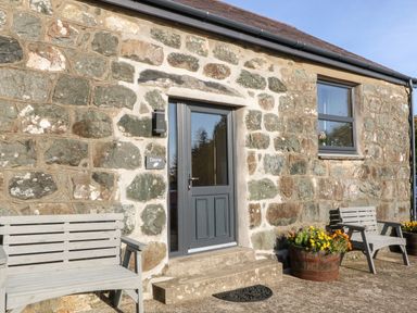 Stone exterior wall with a dark gray door two wooden benches and flower pots at Tyddyn Sianel - Derw in Llanystumdwy near Criccieth