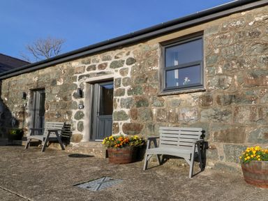 A stone exterior wall with two doors and one window with benches and flower pots outside at Tyddyn Sianel - Derw in Llanystumdwy near Criccieth