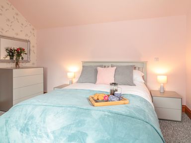 A bedroom with a bed with blue blanket and pillows two bedside tables with lamps and a chest of drawers with flowers at Glenhaven in Trewarmett near Tintagel