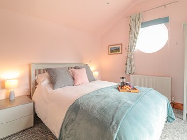 A bedroom with a bed with pillows and a blanket a nightstand with a lamp and a round window with curtains at Glenhaven in Trewarmett near Tintagel