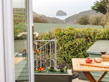 A view of a garden with a hedge and hills beyond with a rock in the sea seen through an open door with a wooden table holding bread and a glass of wine at Glenhaven in Trewarmett near Tintagel