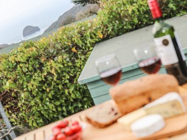 A table outdoors with two glasses of red wine bread cheese and tomatoes with a hedge and coastal view in the background at Glenhaven in Trewarmett near Tintagel