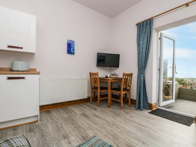 A small dining area with a wooden table and two chairs a wall-mounted television a kitchen counter with a toaster wooden floor and a glass door leading to an outdoor patio at Glenhaven in Trewarmett near Tintagel