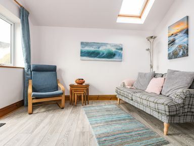 A sitting room with a plaid sofa, blue armchair, nested wooden tables with fruit bowl, and ocean artwork on the walls at Glenhaven in Trewarmett near Tintagel