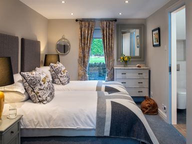 A bedroom with two single beds with patterned pillows a dresser with a mirror yellow flowers and a door to a bathroom at Oakdene Country House in Sedbergh