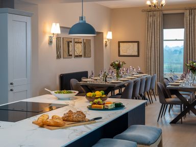 A dining room with a long table set for a meal and a kitchen island with bread salad and cheese at Oakdene Country House in Sedbergh