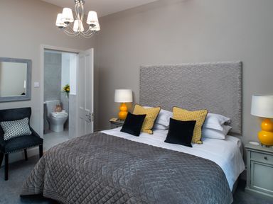 A bedroom with a bed with yellow and black pillows two bedside tables with lamps a chair and a bathroom visible through an open door at Oakdene Country House in Sedbergh