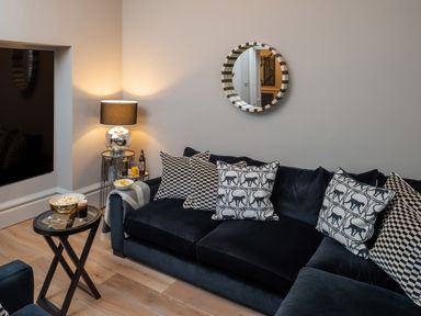 A living room with a dark sofa and patterned cushions a round mirror and a lamp on a side table at Oakdene Country House in Sedbergh