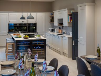 A kitchen with white cabinets an island with stools wine coolers and dining table with place settings at Oakdene Country House in Sedbergh