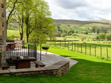 An outdoor patio with a wooden table and benches a hot tub and a fire pit overlooking a fenced green field and hills at Oakdene Country House in Sedbergh