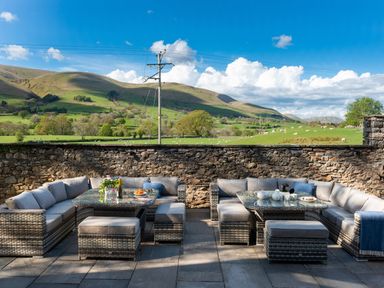 An outdoor seating area with cushioned wicker sofas and tables set on a stone patio with hills and fields in the background at Oakdene Country House in Sedbergh