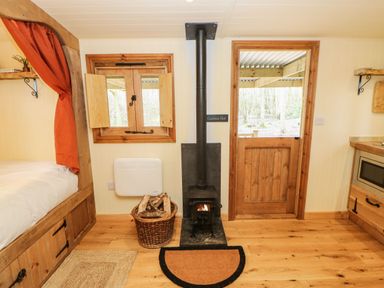 A small room with a wood-burning stove a basket of firewood a wooden door and window and part of a bed at Bracken Fern in Eppleby