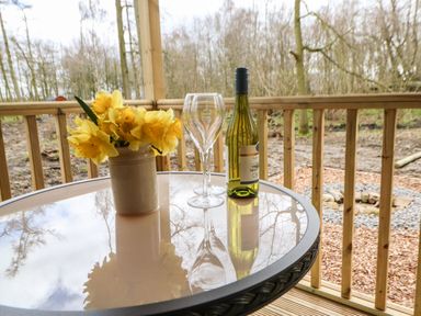 A glass table on a wooden deck with a vase of yellow flowers two wine glasses and a bottle of wine surrounded by trees at Bracken Fern in Eppleby