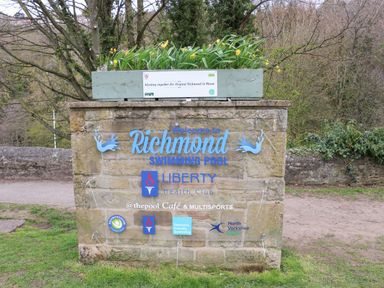A stone sign with information about Richmond Swimming Pool and sponsors with a planter box on top with flowers at Bracken Fern in Eppleby