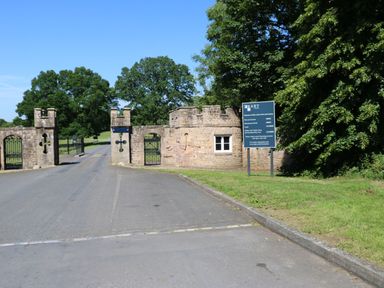 Stone entrance gate with iron gates and a signpost by a roadside with trees in the background at Bracken Fern in Eppleby