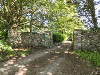 An open gate with stone walls leading to a driveway lined with trees and bushes at Culdrose Manor in Helston