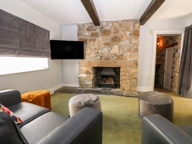A living room with a stone fireplace a wall-mounted TV armchairs and ottomans at Culdrose Manor in Helston