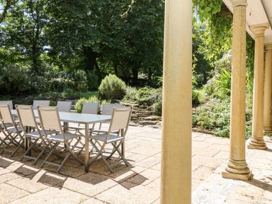 An outdoor patio with a long dining table and chairs surrounded by stone columns and garden greenery at Culdrose Manor in Helston