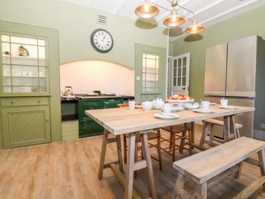 A kitchen with a wooden dining table set with white dishes and cups a green stove built into the wall a clock above it and a large silver refrigerator at Culdrose Manor Helston
