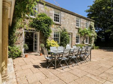 An outdoor patio with a long dining table and chairs in front of a stone building with windows and plants at Culdrose Manor in Helston