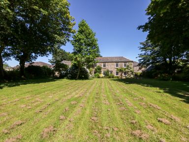 A large grassy yard with trees surrounding a stone house under a clear sky at Culdrose Manor in Helston