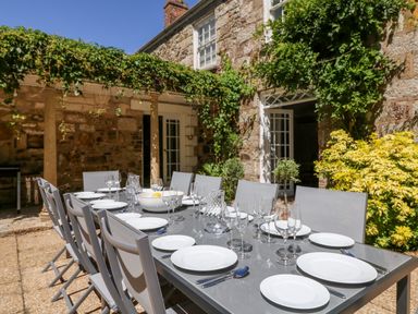 An outdoor dining area with a table set with plates glasses and cutlery surrounded by chairs near a stone house with greenery and open doors at Culdrose Manor in Helston