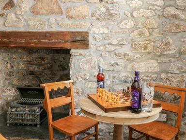 A small round wooden table with a chess board and two bottles of drink with glasses between two wooden chairs in front of a stone wall and a fireplace at Culdrose Manor in Helston