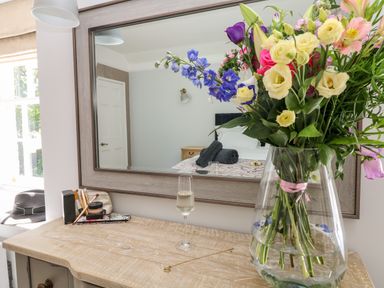 A wooden dresser with a vase of flowers a glass of drink and makeup items in front of a mirror reflecting a bed and door at Culdrose Manor in Helston