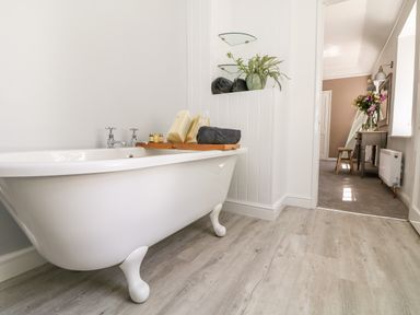 A bathroom with a white freestanding bathtub with wooden floor and a hallway view with a table and flowers at Culdrose Manor in Helston