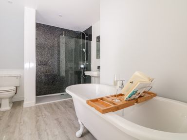 A bathroom with a white clawfoot bathtub holding a wooden tray with a glass and a book a black tiled shower a white toilet and a sink at Culdrose Manor in Helston
