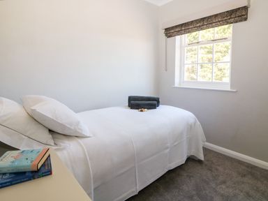 A bedroom with a single bed covered with white bedding and pillows a window with a blind and books on a bedside table at Culdrose Manor in Helston