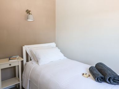A bedroom with a single bed white linens two rolled towels on the bed a small bedside table with books and a wall light above the bed at Culdrose Manor in Helston