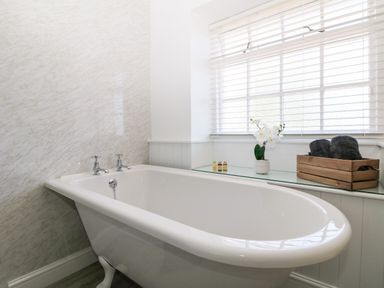 A bathroom with a white freestanding bathtub under a window with blinds and a shelf holding a wooden crate and small bottles at Culdrose Manor in Helston