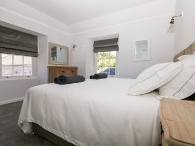 A bedroom with a bed covered in white bedding two rolled towels on the bed two windows with grey blinds a wooden chest of drawers and two wall mirrors at Culdrose Manor Helston