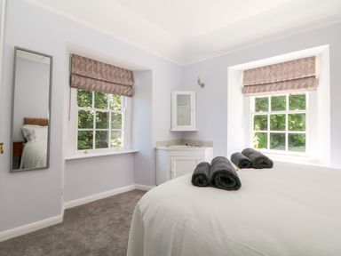 A bedroom with two windows with patterned blinds a bed with rolled towels on white bedding a wall mirror and a corner sink unit at Culdrose Manor in Helston