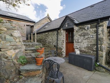 An outdoor stone patio with a metal table and chairs potted plants stone steps and a cat on a black storage box at Nyth Y Wennol in Llanfachraeth