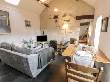 A living room with a grey sofa a wooden dining table with two chairs a green armchair a TV on a stand and a stone wall at Nyth Y Wennol in Llanfachraeth