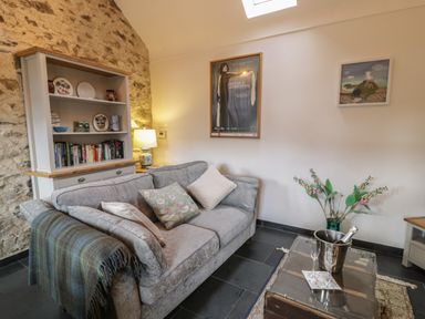 A living room with a grey sofa cushions a throw blanket a bookshelf a table with champagne and glasses flowers and framed pictures on the wall at Nyth Y Wennol in Llanfachraeth