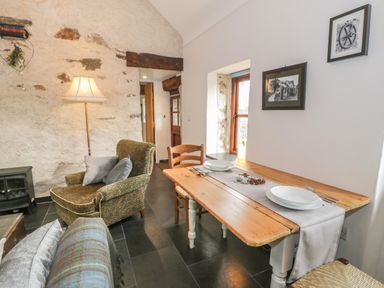 A small dining area with a wooden table set for two and an armchair next to a lamp in a room with stone walls at Nyth Y Wennol in Llanfachraeth