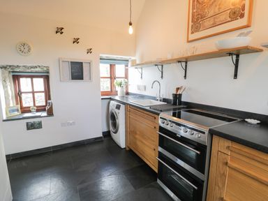 A kitchen with wooden cabinets a washing machine a stove two windows and shelves with dishes at Nyth Y Wennol in Llanfachraeth