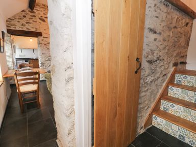A narrow dining area with a wooden table and chairs next to a stone wall and stairs with patterned tiles at Nyth Y Wennol in Llanfachraeth