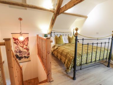 A bedroom with a metal bed with gold and black frame and patterned yellow bedspread near wooden stairs with butterfly wall hanging at Nyth Y Wennol in Llanfachraeth