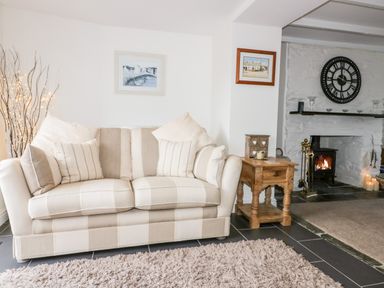 A living room with a beige striped sofa wooden side table and a fireplace with a large clock on the wall at Hawthorn Cottage in St Teath