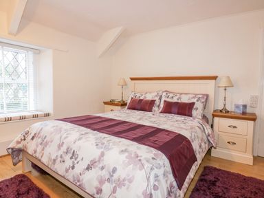 A bedroom with a double bed floral bedding purple accents two bedside tables lamps and a window with blinds at Hawthorn Cottage in St Teath