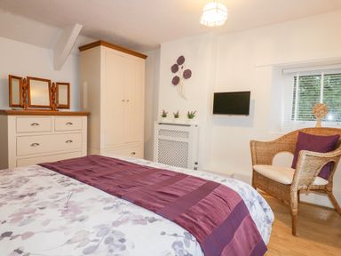 A bedroom with a bed covered with patterned and purple bedding a wicker chair with a purple cushion a chest of drawers with mirrors a wardrobe a wall-mounted TV and small plants on a radiator cover at Hawthorn Cottage in St Teath